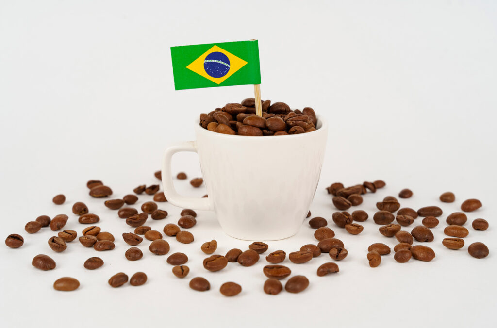 Grãos de café e uma bandeira do Brasil dentro de uma xícara. Também há grãos de café em volta da xícara. A imagem simboliza o poder da exportação de café do Brasil.